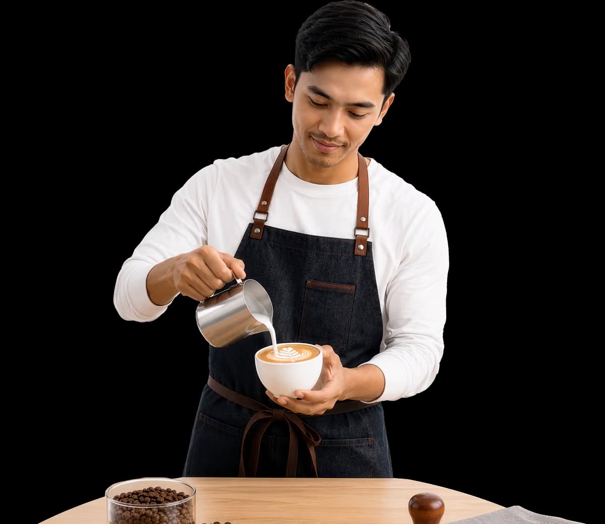 Barista in apron pouring milk into a cup of coffee with latte art on a wooden table.