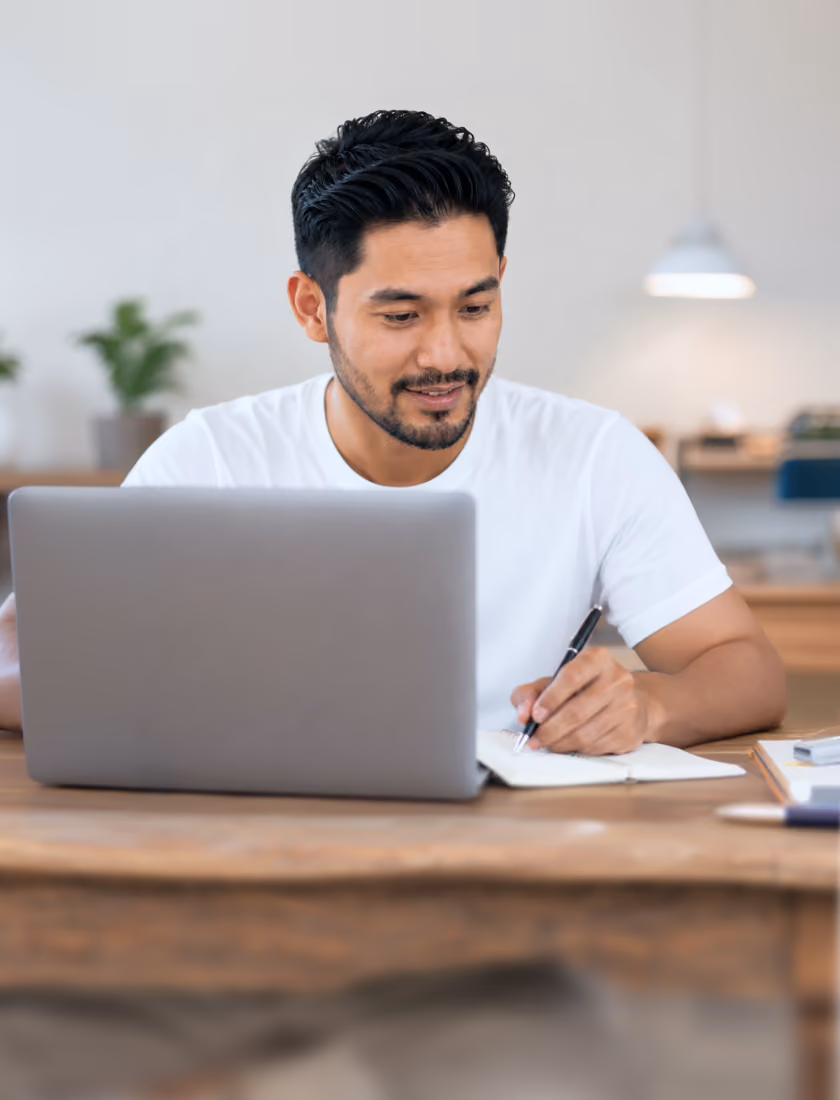 A man writing notes on paper in front of his laptop.